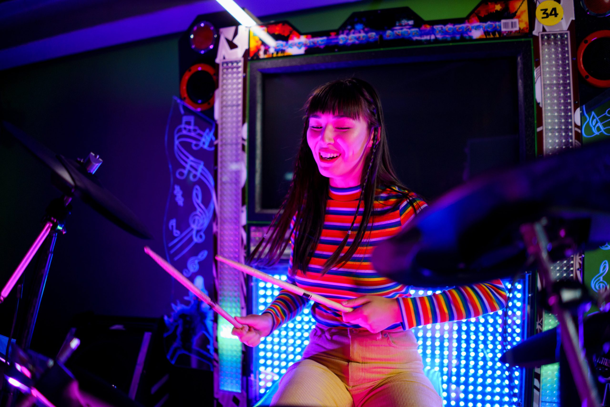 Une jeune femme d'origine asisatique jouant de la batterie, devant un écran et des petites lumières bleues
